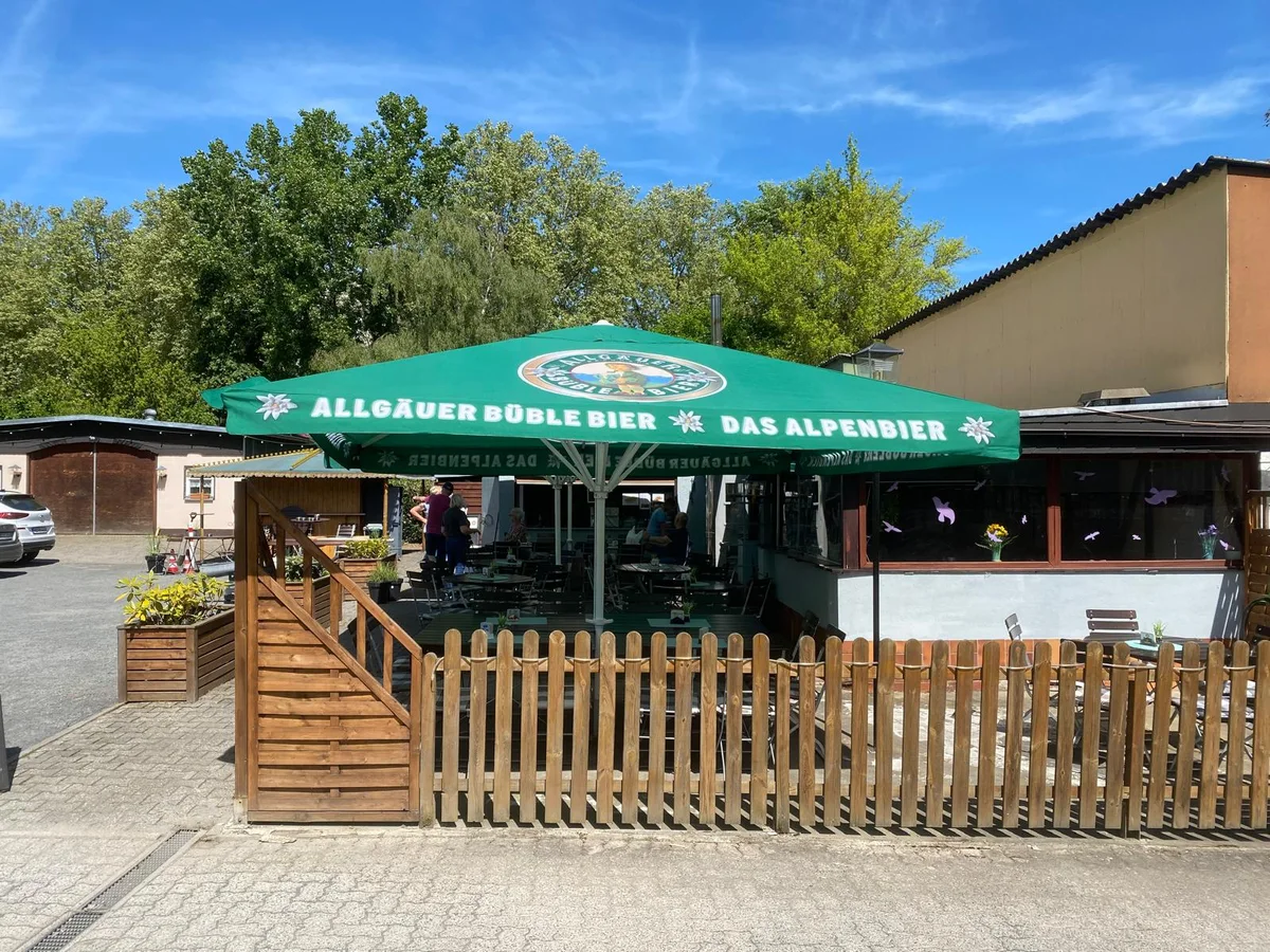 Vereinslokal mit Holzzaun und Tischen unter einem großen grünen Sonnenschirm